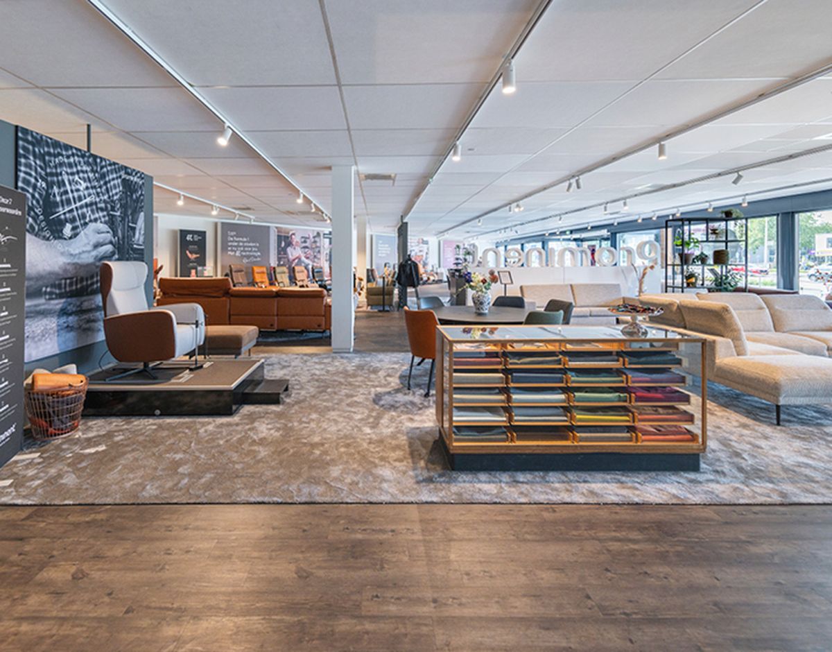 Meubelwinkelinterieur met moderne banken en stoelen, een rek met stoffenstalen op de voorgrond, en grote ramen die veel licht binnenlaten.