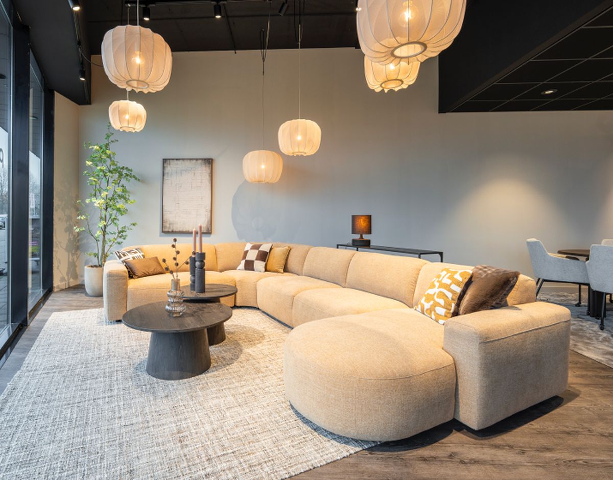 Moderne woonkamer met beige hoekbank, ronde salontafel en hanglampen. Grote ramen zorgen voor veel lichtinval. Decoratieve kussens en plant.