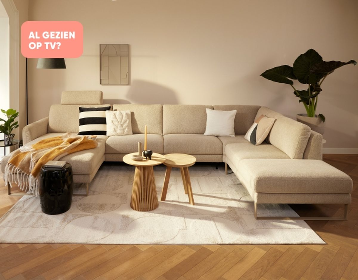 Modern living room with a beige sectional sofa, decorative pillows, round nesting tables, a white rug, and a plant in the corner.