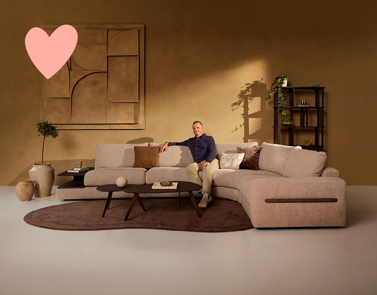 A man sits on a beige sectional sofa in a warmly lit living room with abstract wall art, a round coffee table, plants, and a decorative shelf.