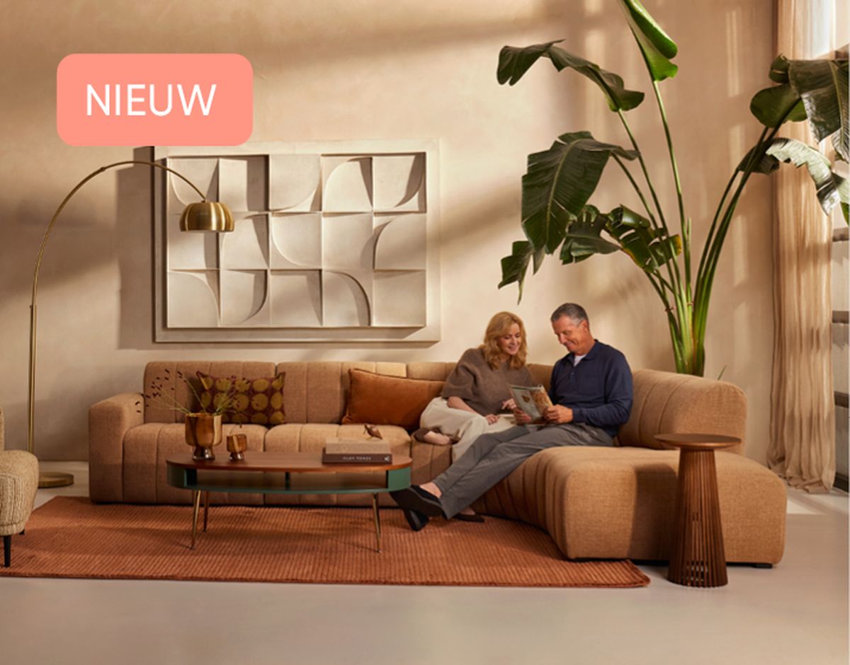 A couple sitting on a modern beige sectional sofa, reading a magazine. A large plant and abstract art decorate the room. "NIEUW" in corner.