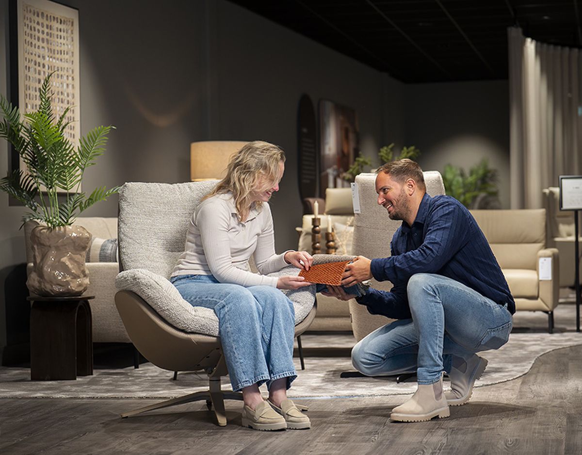 Een man geeft een vrouw een cadeau terwijl ze samen in een meubelwinkel zitten. Ze glimlachen en de ruimte is modern ingericht met planten.
