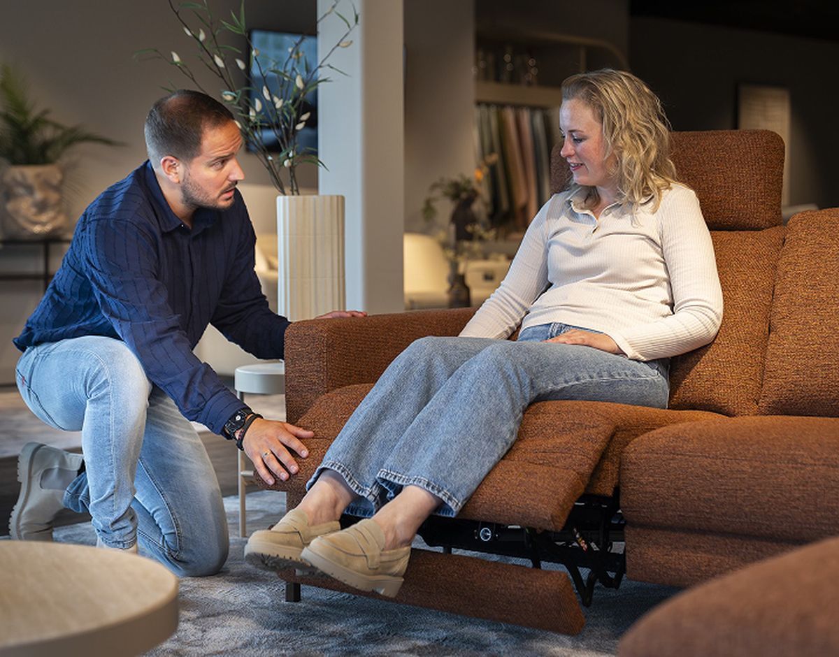 Een vrouw zit op een bruine verstelbare bank met een ontspannen houding, terwijl een man naast haar knielt en met haar praat.
