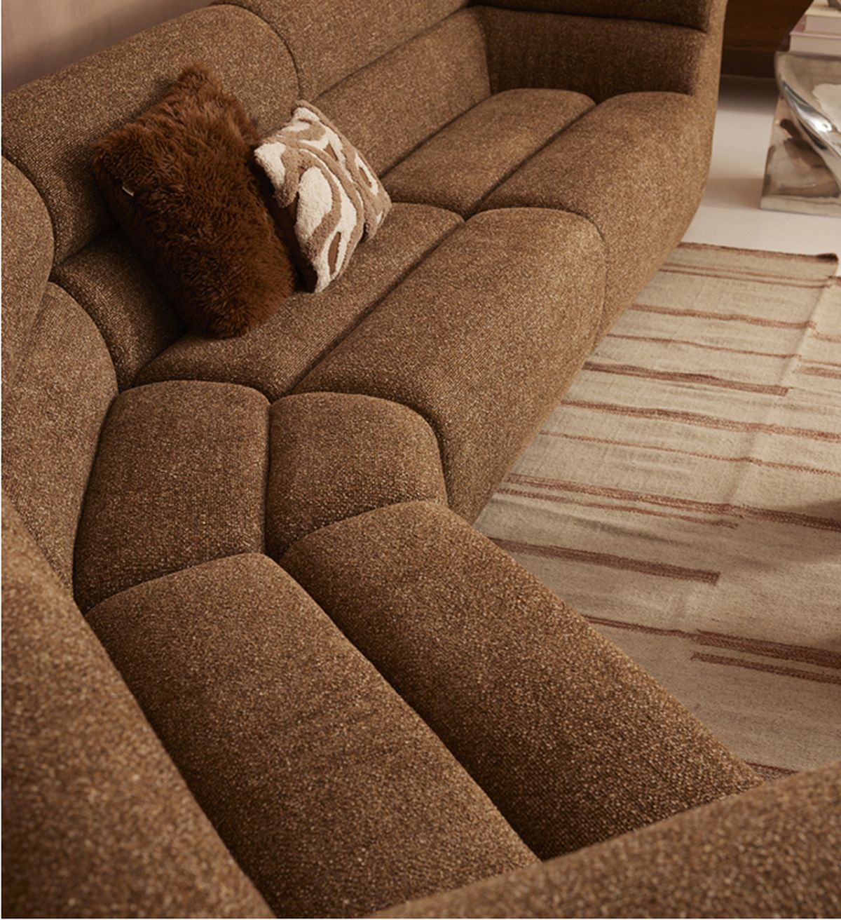 A brown textured sectional sofa with two decorative pillows, one fluffy and one patterned, next to a striped rug on a light floor.