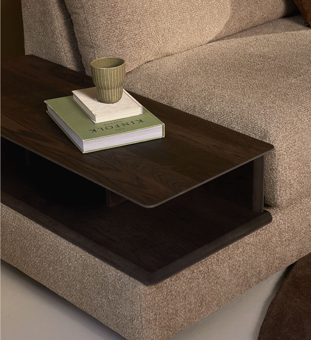 A close-up of a wooden side table attached to a sofa, featuring a green cup and two books on top, with a textured beige fabric cushion.