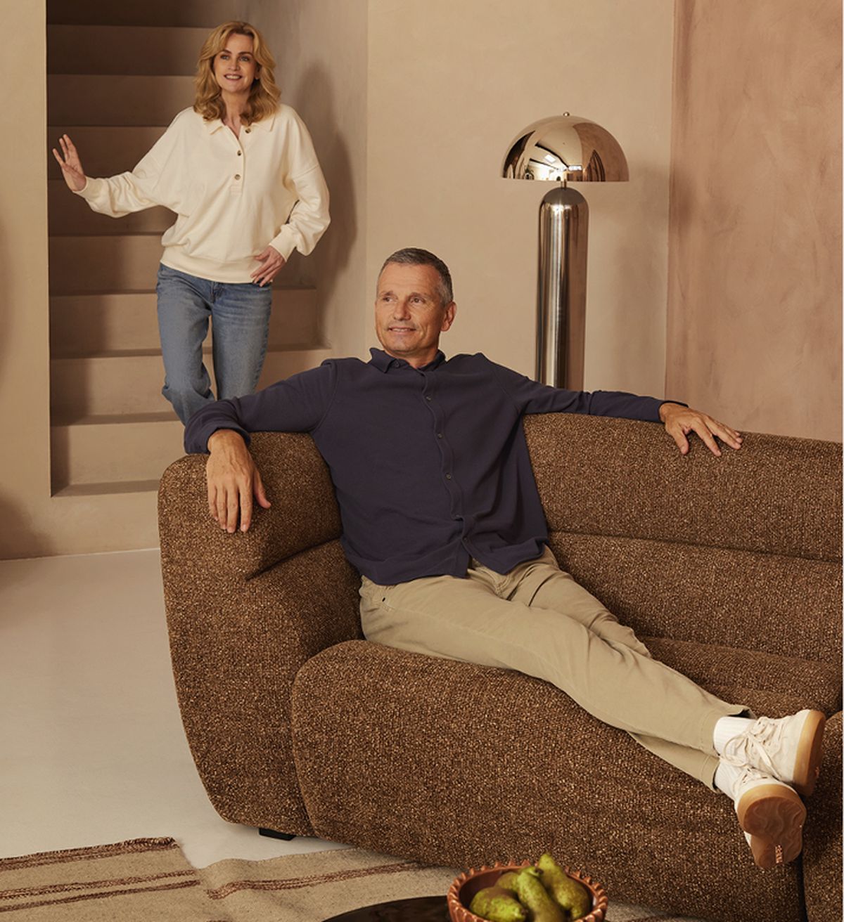 A man sits relaxed on a brown sofa while a woman stands on stairs nearby. A modern lamp and a bowl of pears are seen in the room.
