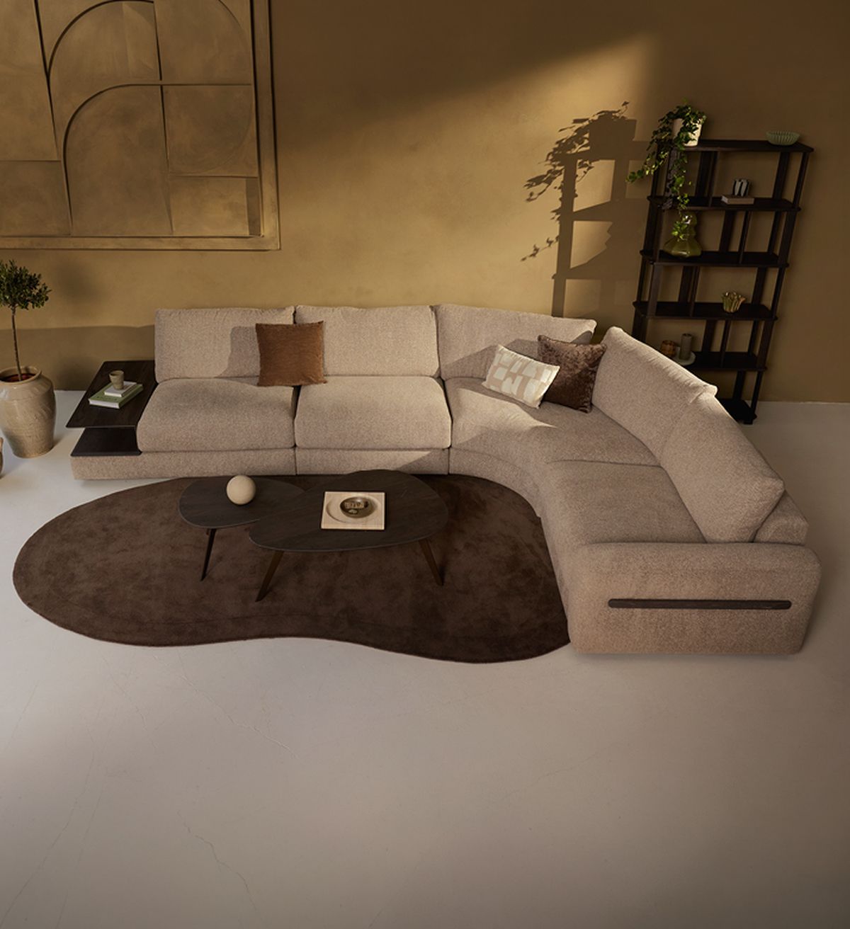 Modern living room with a beige sectional sofa, brown cushions, curved coffee tables on a brown rug, wall art, and a tall shelf with plants.
