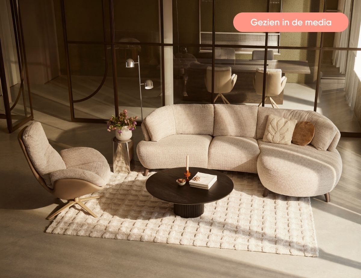 Modern living room with a beige L-shaped sofa, cozy armchair, round black coffee table, and decor on a textured rug, in a glass-partitioned space.