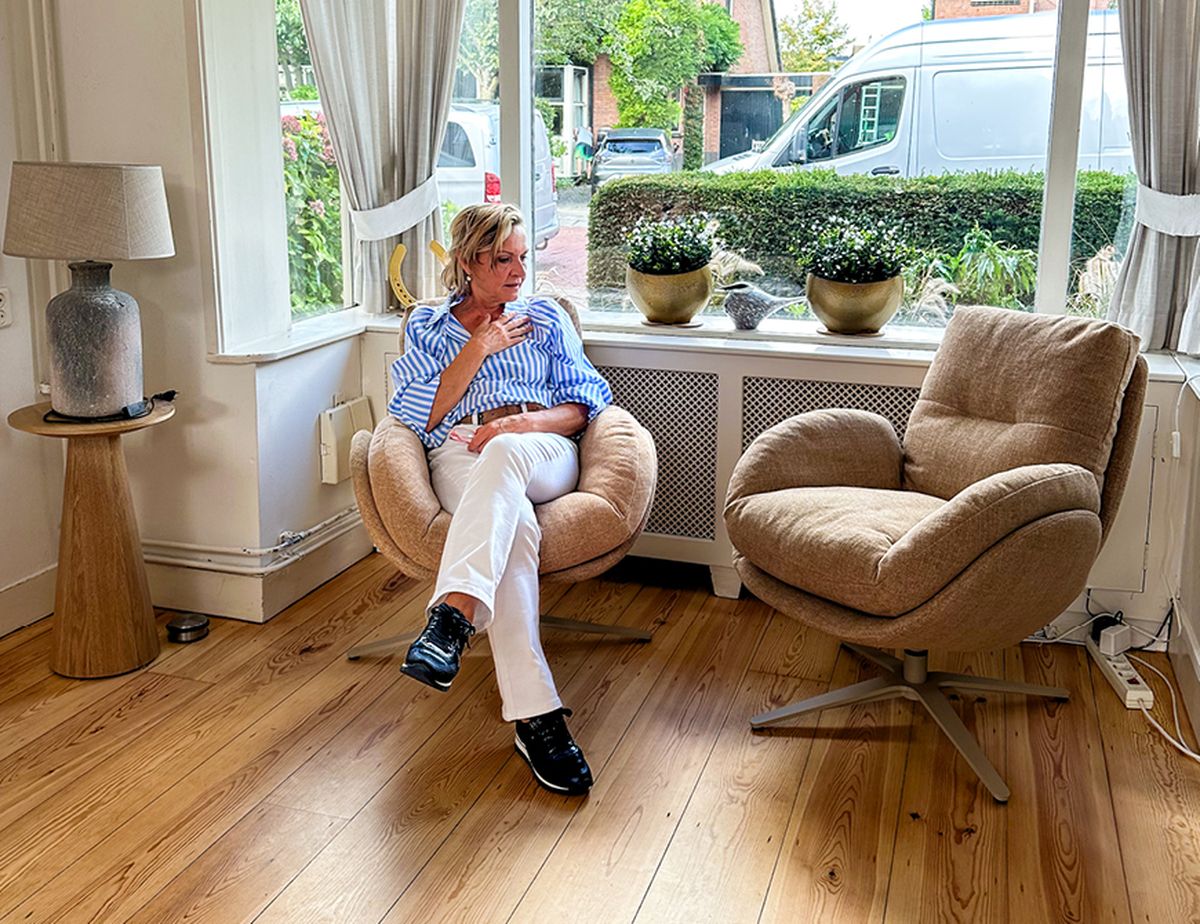 A person in a blue striped shirt and white pants sits in a cozy chair by a window, with two potted plants on the ledge and a lamp on a side table.