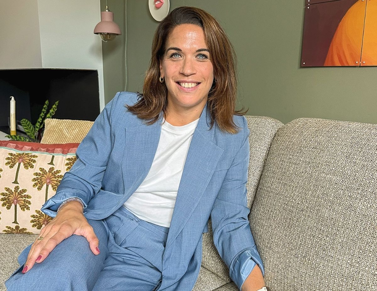 A woman in a blue suit and white shirt sits on a grey sofa, smiling. A decorative pillow and a green plant are visible in the background.