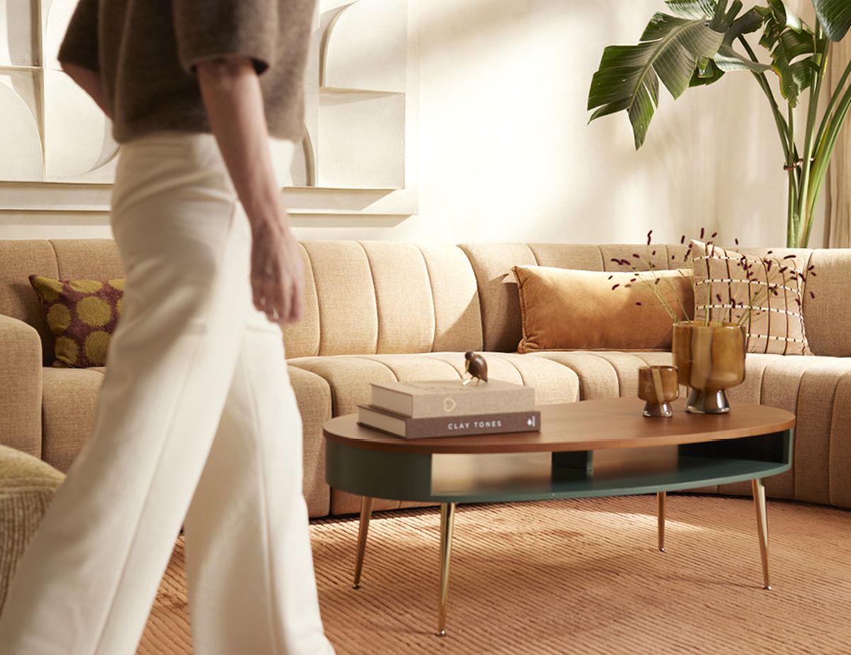 A person in white pants walks past a beige sofa with cushions, a wooden coffee table with books and decor, and a large potted plant in a cozy living room.