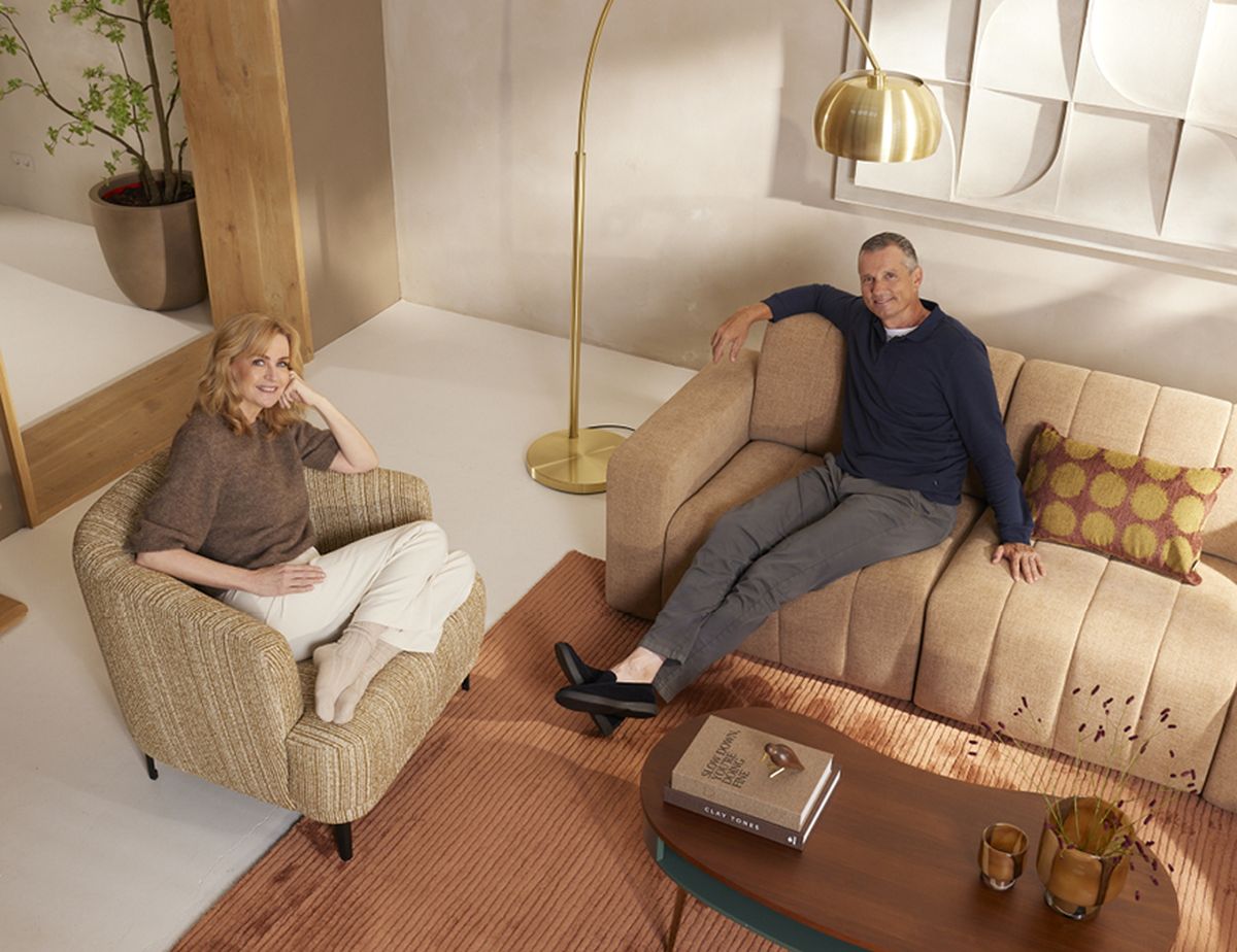 Two people sitting in a modern living room with neutral tones, a floor lamp, a patterned rug, and a coffee table with books and glasses.