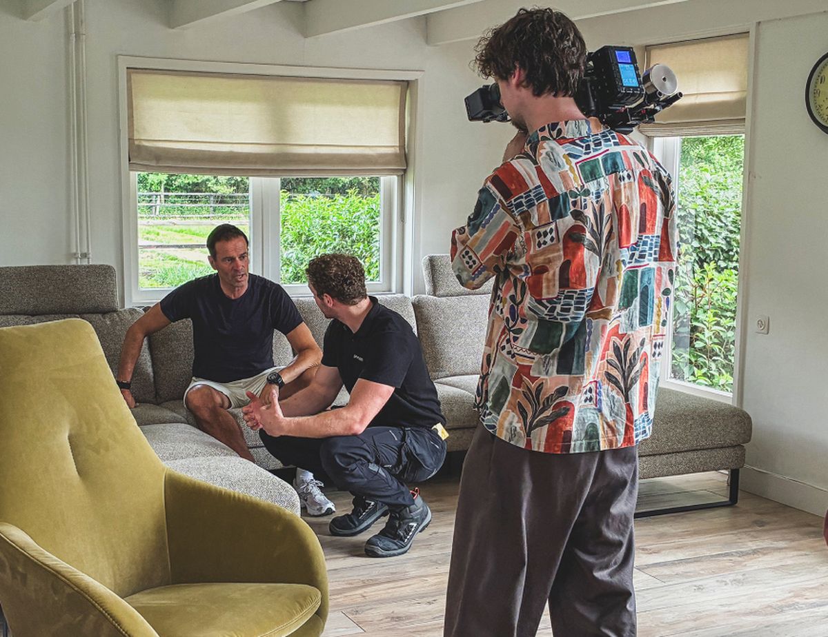 A cameraman films two men sitting on a couch having a conversation in a bright living room with large windows and a yellow chair.