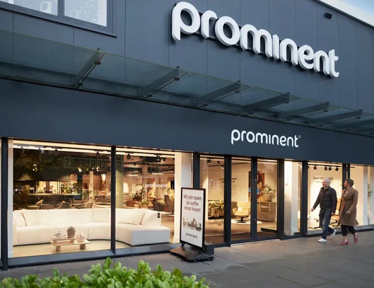 Storefront of Prominent furniture store with large windows displaying a white sofa and a sign outside. Two people walk by on the sidewalk.