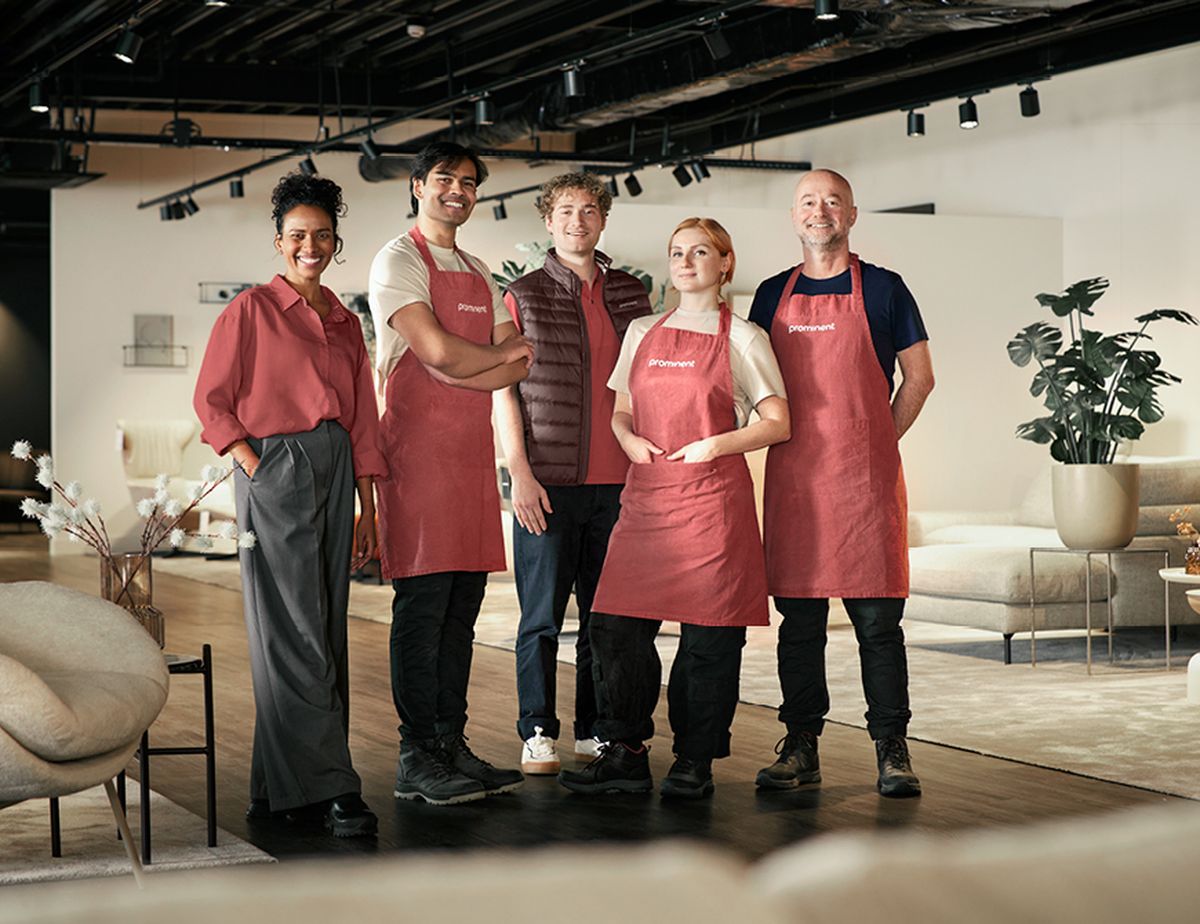 A diverse group of five people stand confidently in a modern furniture showroom, with three wearing matching red aprons.