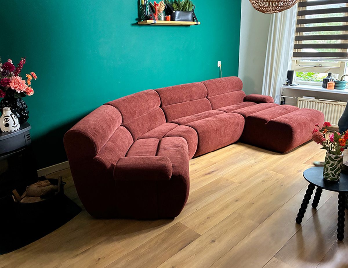 Een gezellige woonkamer met een grote rode hoekbank tegen een groenblauwe muur, een houten vloer, verse bloemen en een raam met jaloezieën.