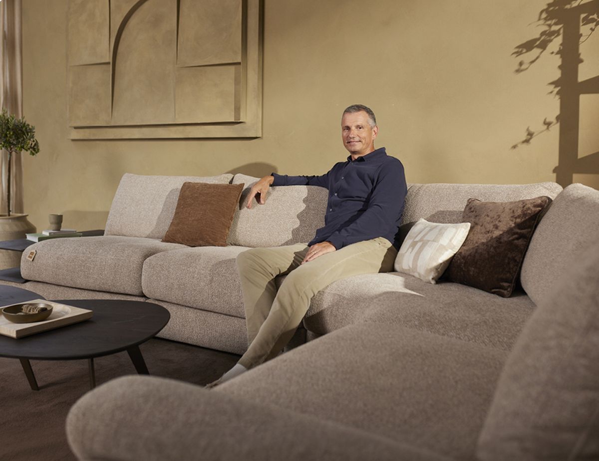 Man sitting on a modern beige sofa in a stylish living room with warm lighting, decorative pillows, and a minimalist wall design.