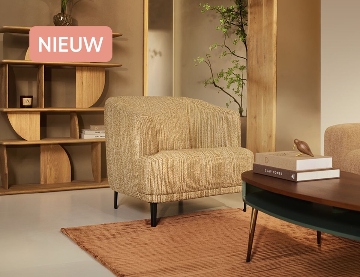 Cozy living room with a beige armchair, wooden coffee table with books, and a bookshelf. A rug is on the floor, and a plant is in the background.
