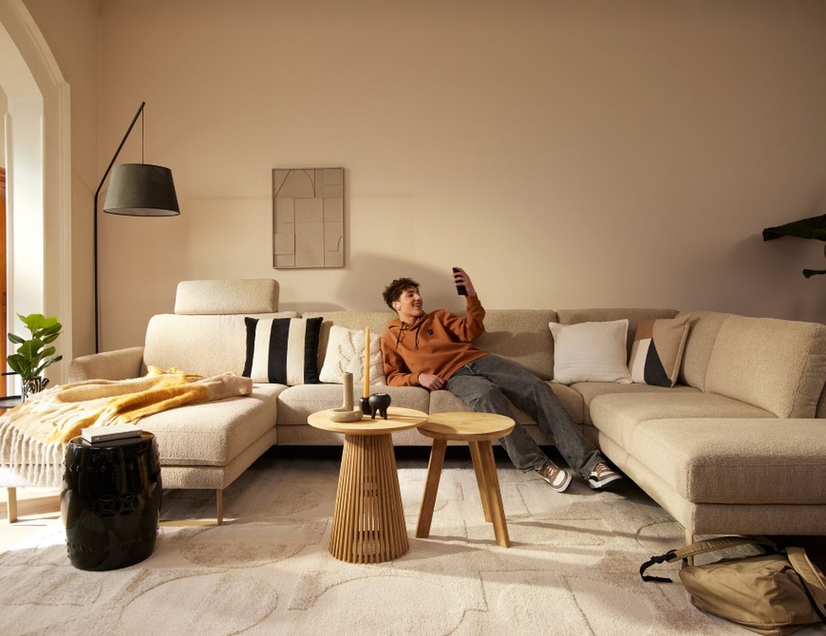 A person in an orange hoodie relaxes on a beige sectional sofa, looking at their phone. The room is stylishly decorated with a modern aesthetic.