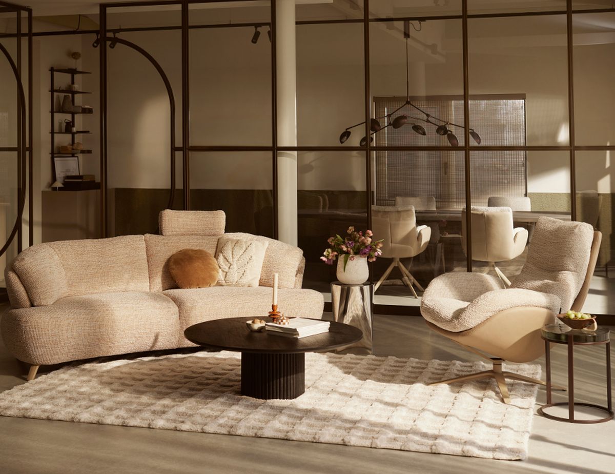 Modern living room with a beige sofa and chair on a textured rug, a round coffee table, and a vase of flowers, with glass partitions in the background.