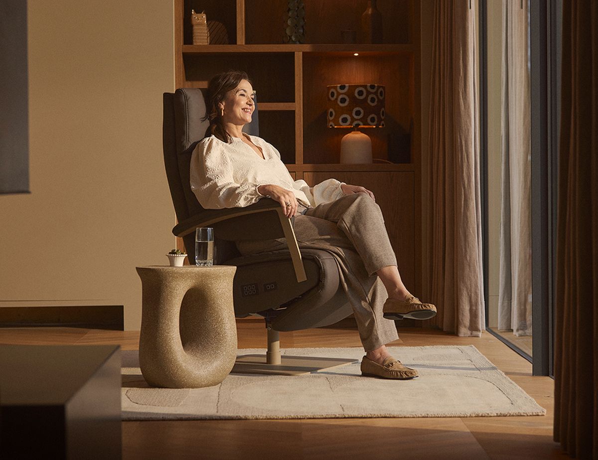 Woman relaxing in a reclining chair by a window, with a side table and a glass of water nearby, in a warmly lit room with shelves in the background.