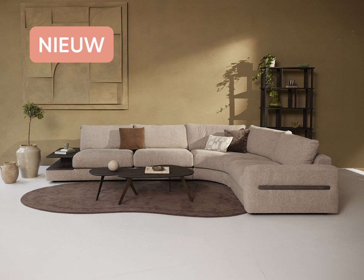 Moderne woonkamer met een beige L-vormige bank, een bruin vloerkleed, een salontafel, planten en ingebouwde planken tegen een zandkleurige muur. Label 'NIEUW' linksboven.