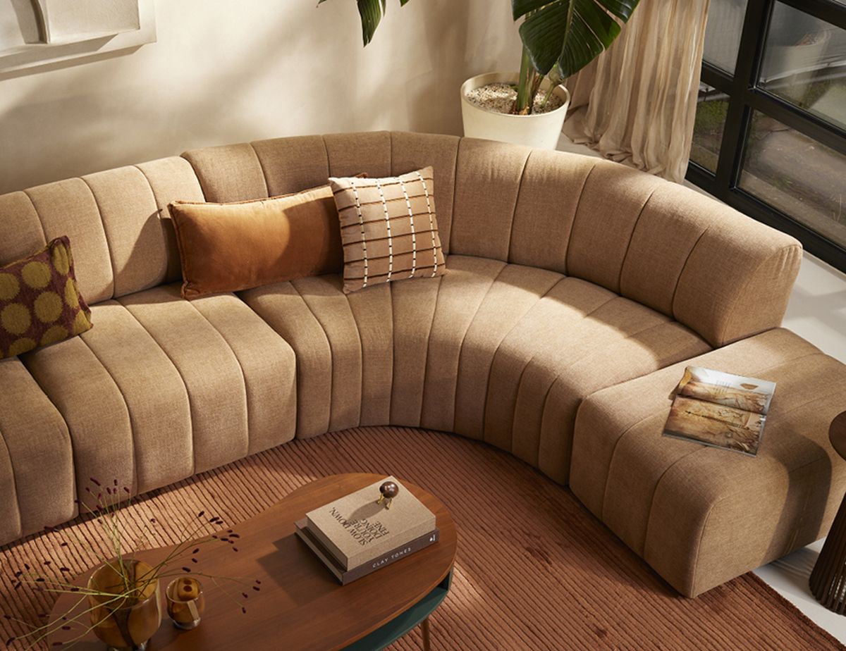 Curved beige sofa with two decorative pillows in a cozy living room, plant in corner, books and decorations on wooden coffee table, large window nearby.