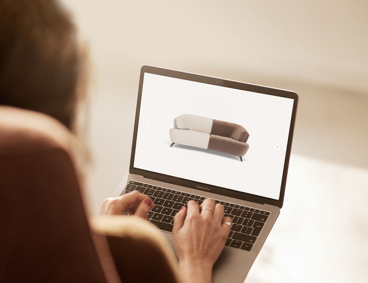 Person using a laptop, viewing a two-toned sofa on the screen in a bright setting.