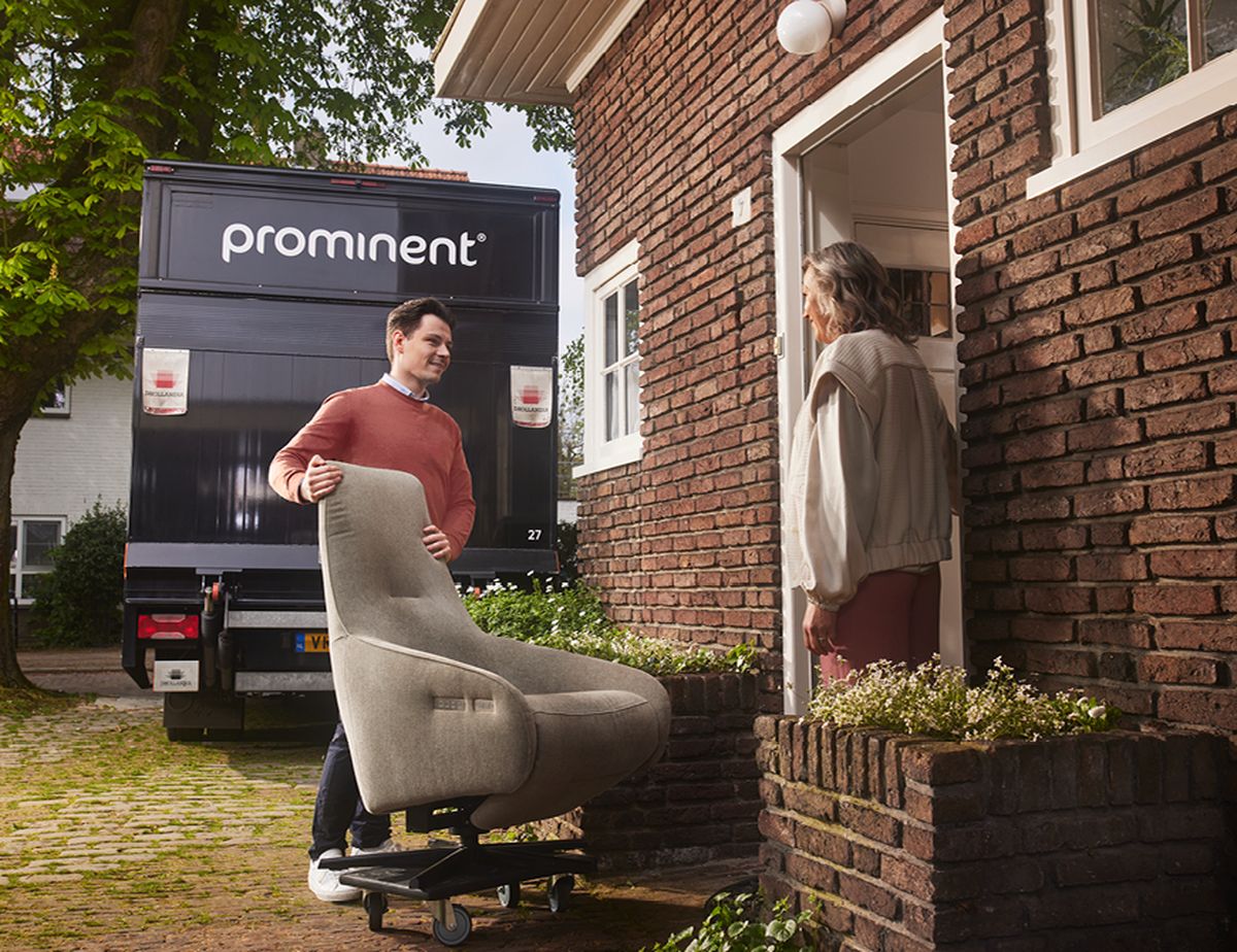 Een man levert een fauteuil af bij de voordeur van een huis, met een vrachtwagen met opschrift "prominent" op de achtergrond.