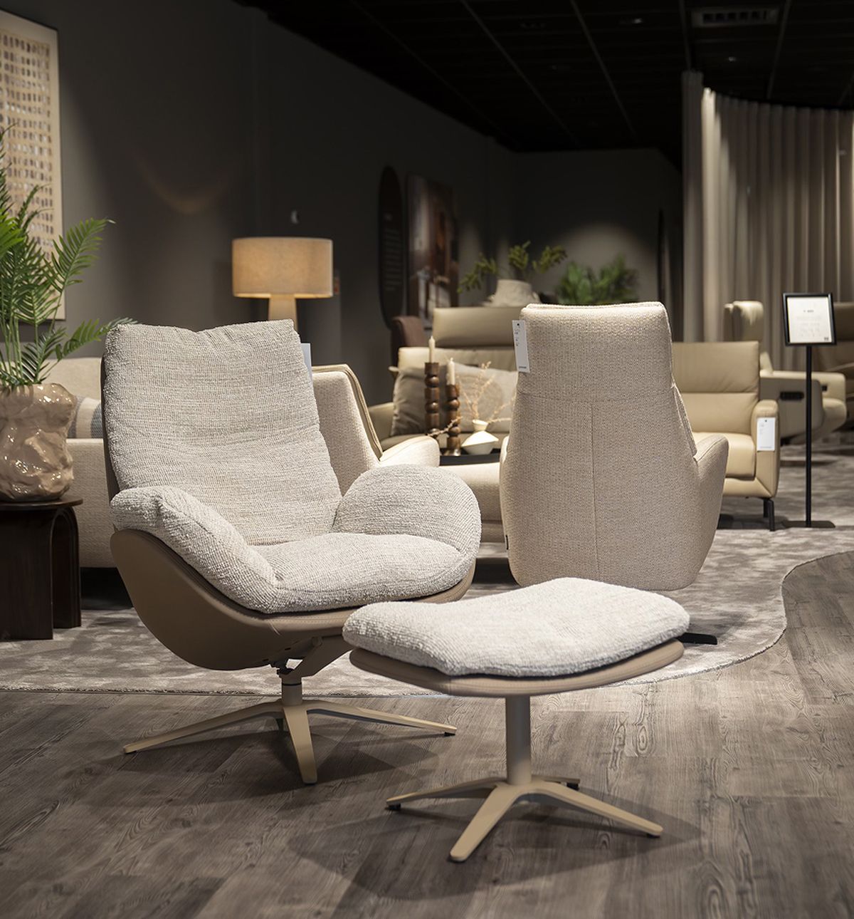 A modern furniture showroom with two armchairs and an ottoman, wooden floor, plants, and warm lighting.