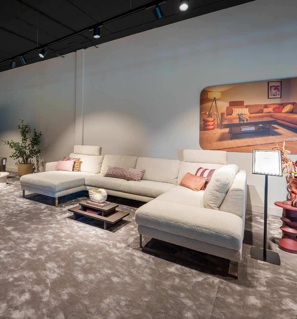 Modern living room with a large, L-shaped cream sofa, colorful cushions, a low coffee table, potted plant, and framed artwork on the wall.