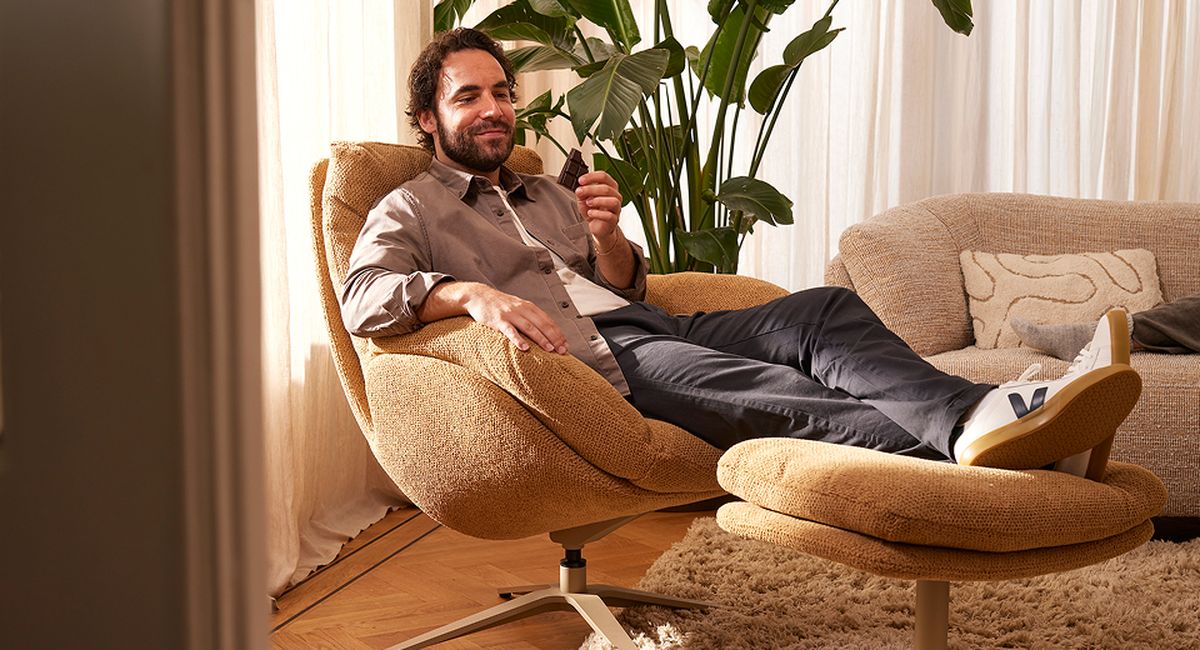 Man ontspant in bruine fauteuil met voetensteun, glimlachend met smartphone in hand. Achtergrond heeft planten en gordijnen.
