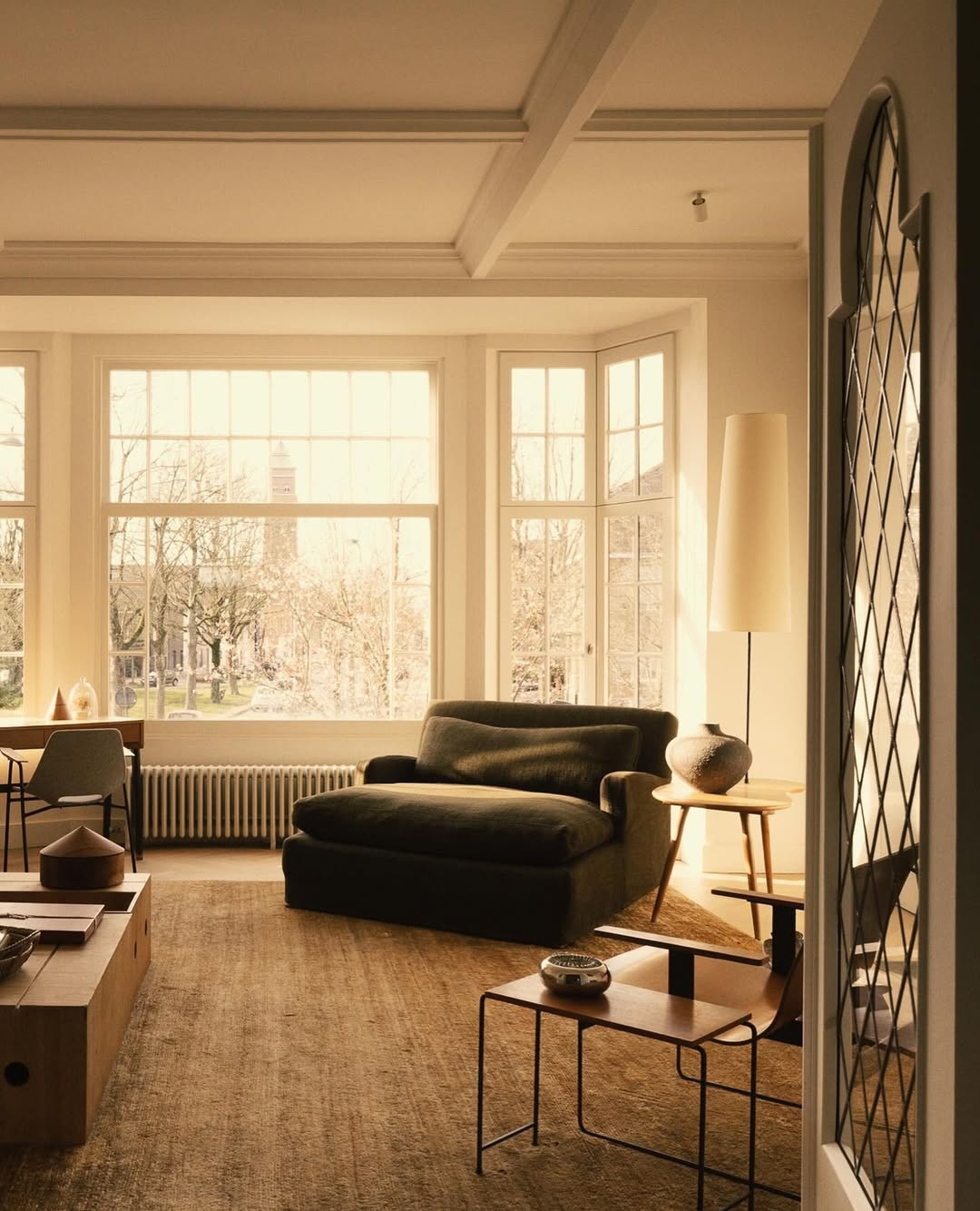 A cozy and inviting living room with natural light streaming through large windows. In the foreground, an Olivia chaise longue with two arms sits on a rustic, woven rug. The space features minimalist furniture, including a modern coffee table and a floor lamp, creating a warm and serene atmosphere.