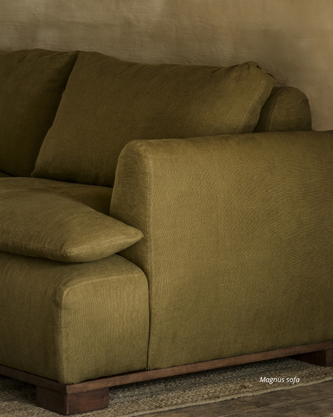 Olive green sofa with plush cushions and wooden base, set against a textured beige wall.