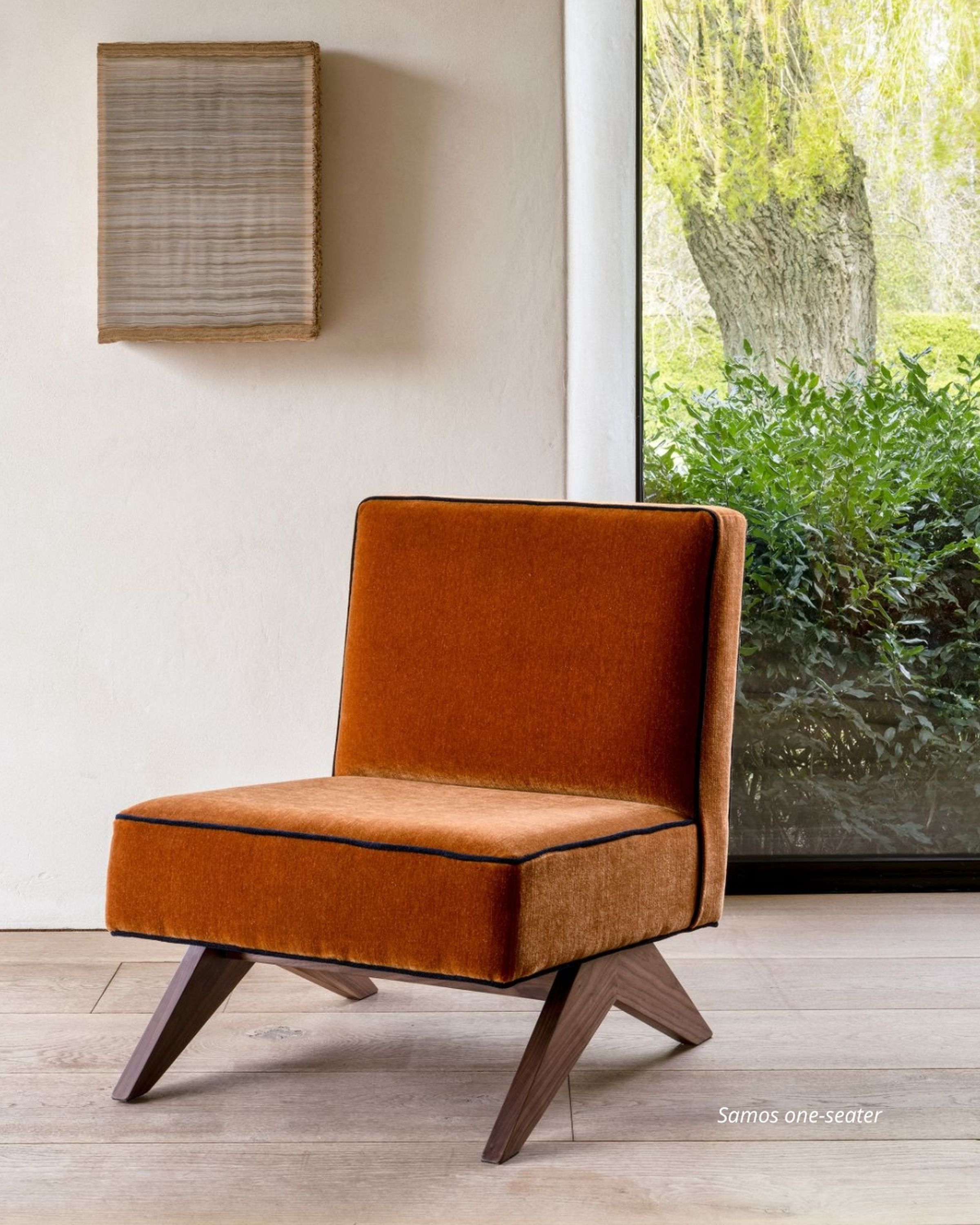 Orange velvet chair with wooden legs on a light wood floor, near a large window with green foliage outside. Wall art hangs above the chair.