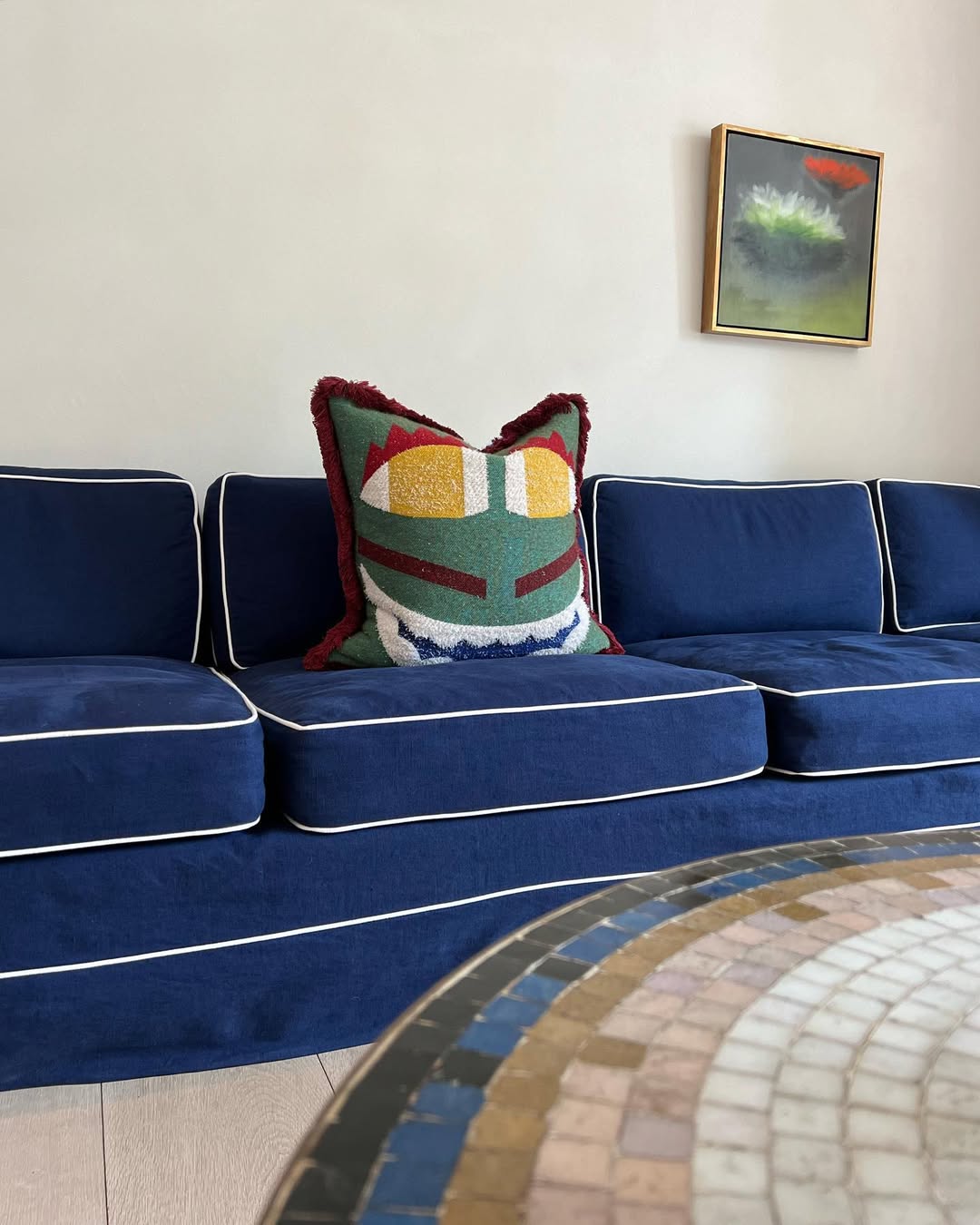 Close-up of a bold royal blue sofa with white piping, styled with a colorful, textured cushion in geometric patterns of green, yellow, red, and white. Above, a small framed abstract artwork adds balance, while the mosaic-tiled tabletop in the foreground introduces earthy tones to the vibrant, modern setting.