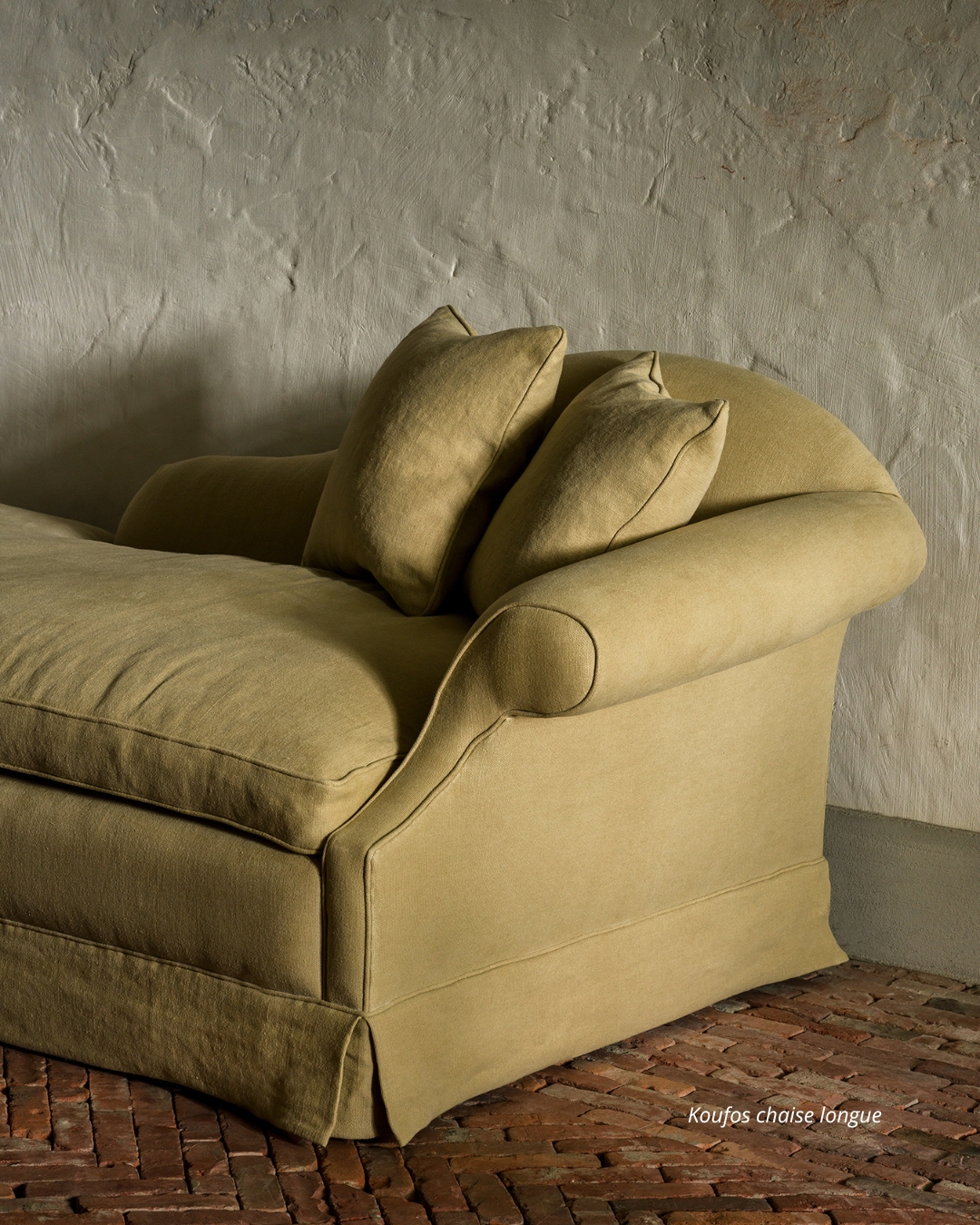 Koufos chaise longue in a soft green fabric with rounded armrests and matching cushions, styled against a textured plaster wall and rustic brick floor.