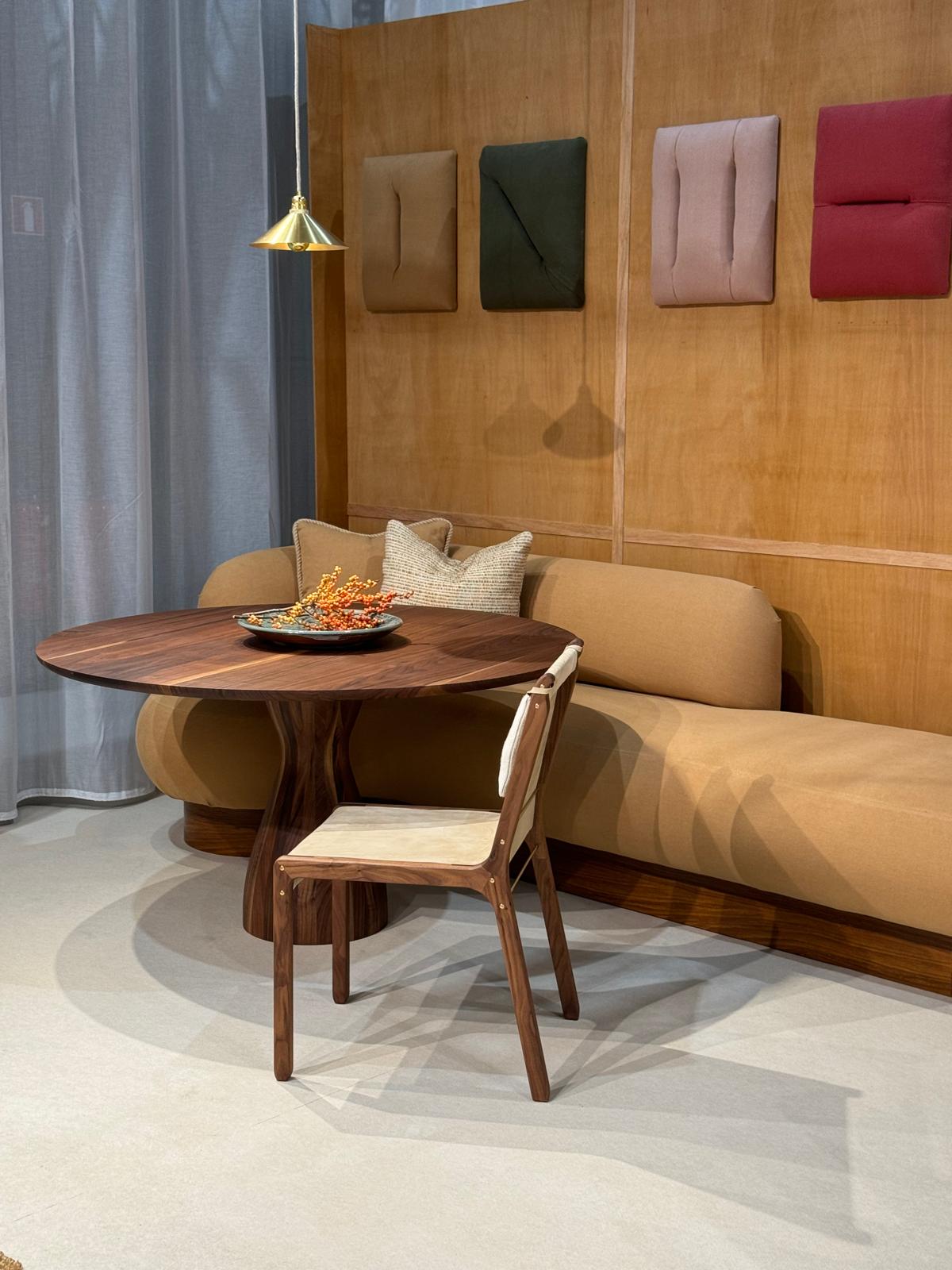 Interior setting with the Nova dining table in walnut wood paired with a matching chair featuring light upholstery, placed next to a curved beige banquette. A brass pendant light hangs above, with acoustic panels in muted colors on the wooden wall behind.