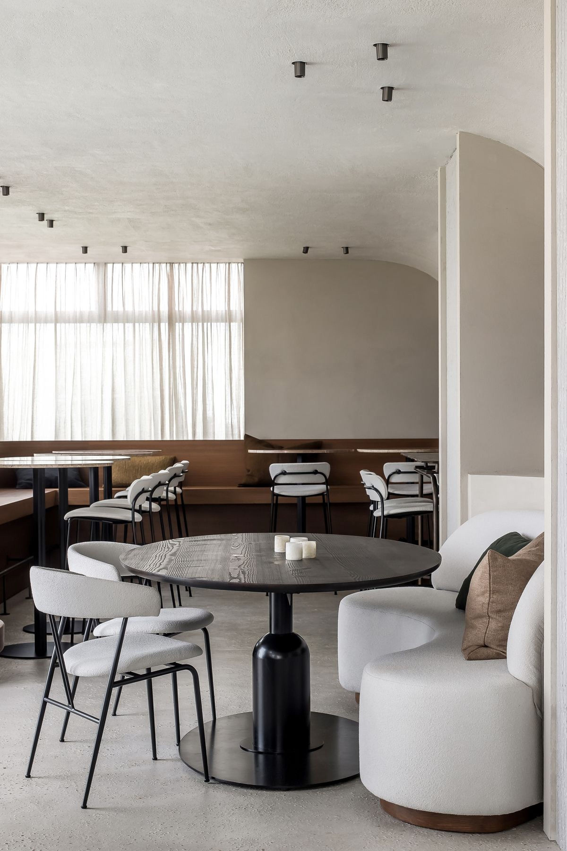 Contemporary restaurant interior featuring a round black table, light upholstered banquette seating with accent pillows, and minimalist design elements under soft natural light.