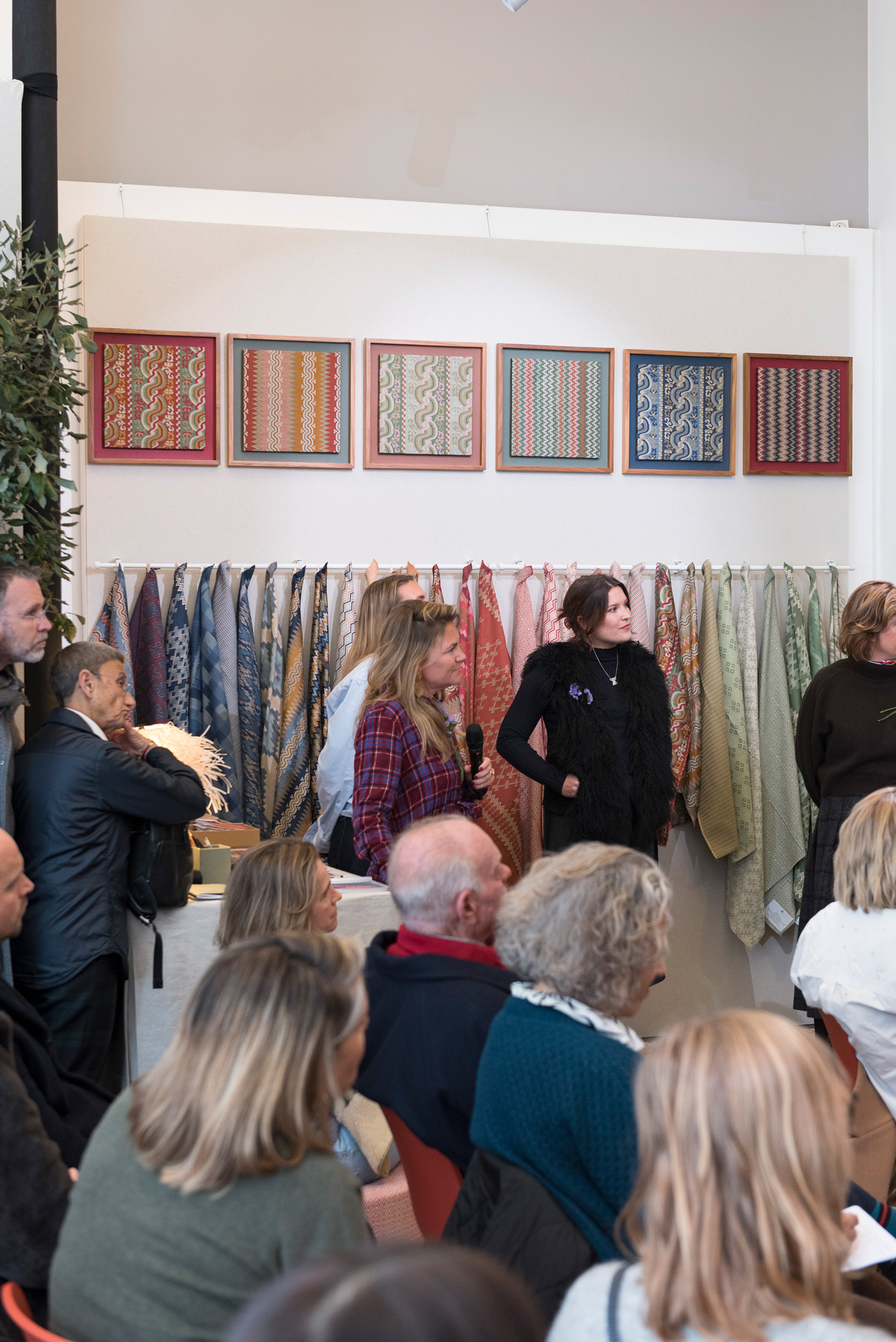 Audience attending a design event with fabric samples and framed textile patterns on display, featuring colorful woven designs in red, blue, green, and beige tones.