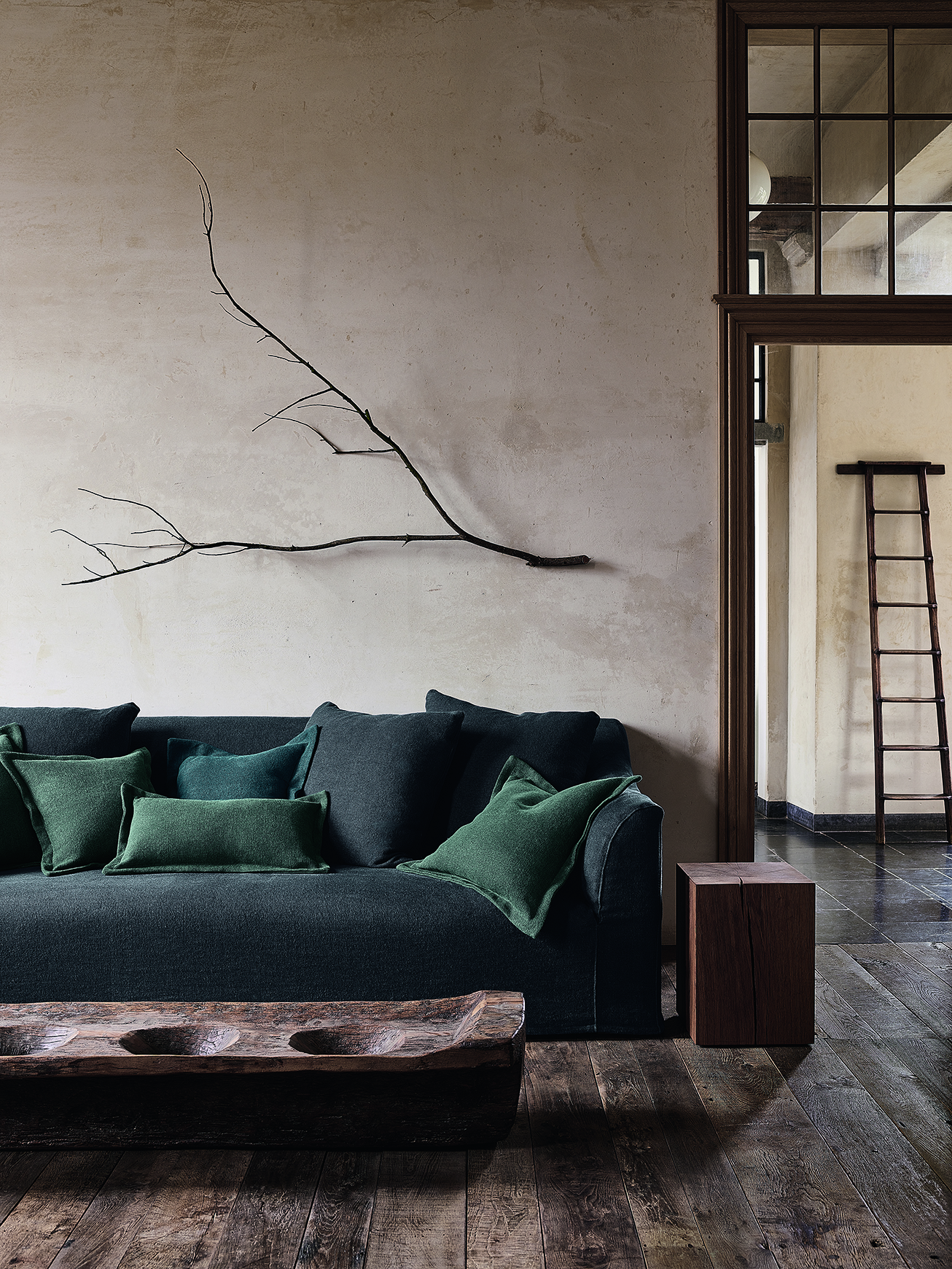 Moody interior with the Toscana fixed seat sofa by Collett & Victor in deep green upholstery, styled with layered cushions and paired with rustic wooden furniture and minimalist wall art.