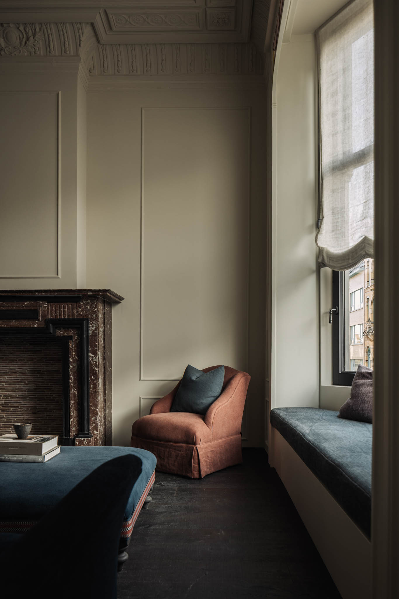 Cozy room with a red armchair, dark fireplace, large window with sheer curtains, and a cushioned window seat, creating a serene atmosphere.
