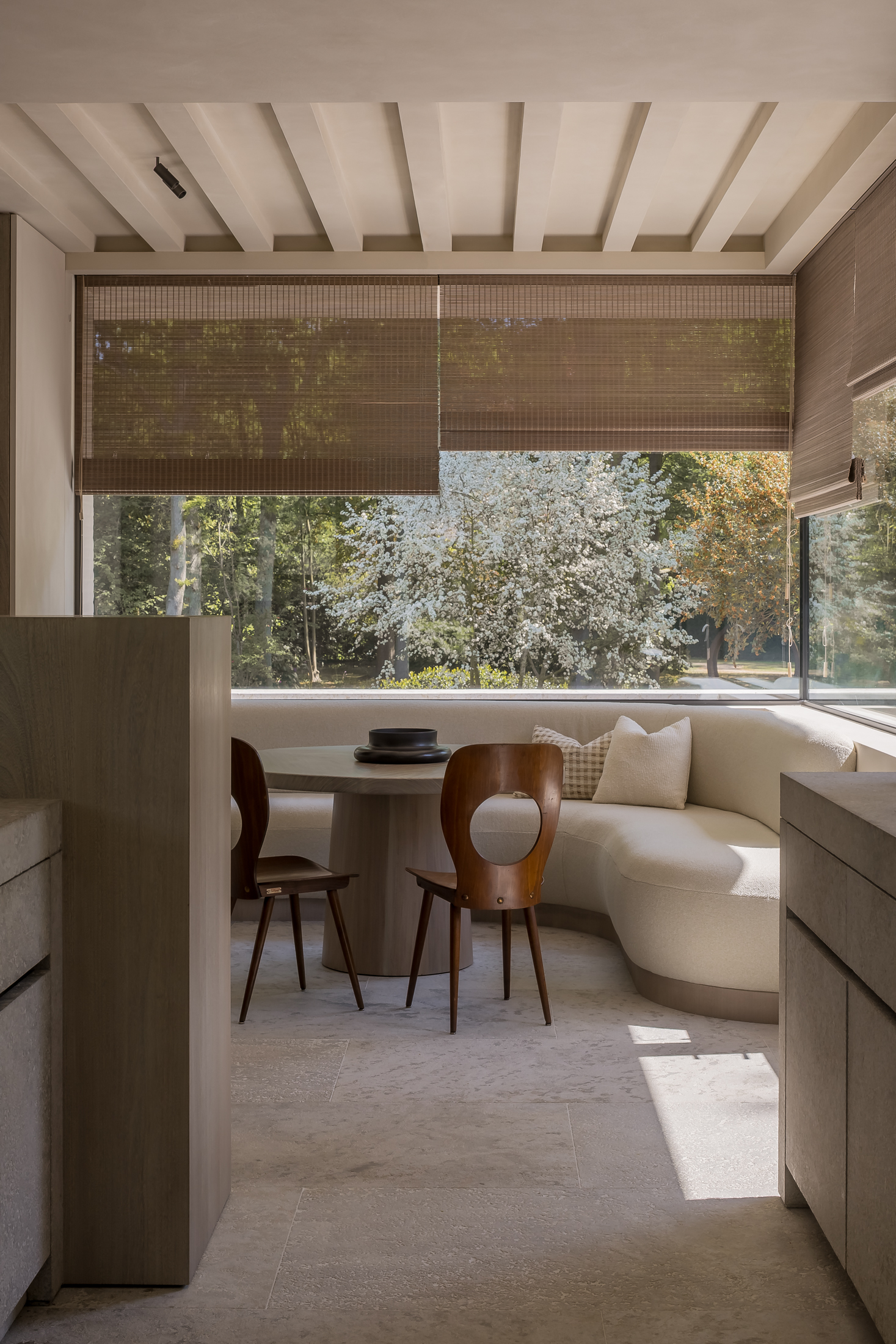 Cozy dining nook with a curved white sofa, two wooden chairs, and a round table. Large windows with blinds offer a view of lush greenery outside.