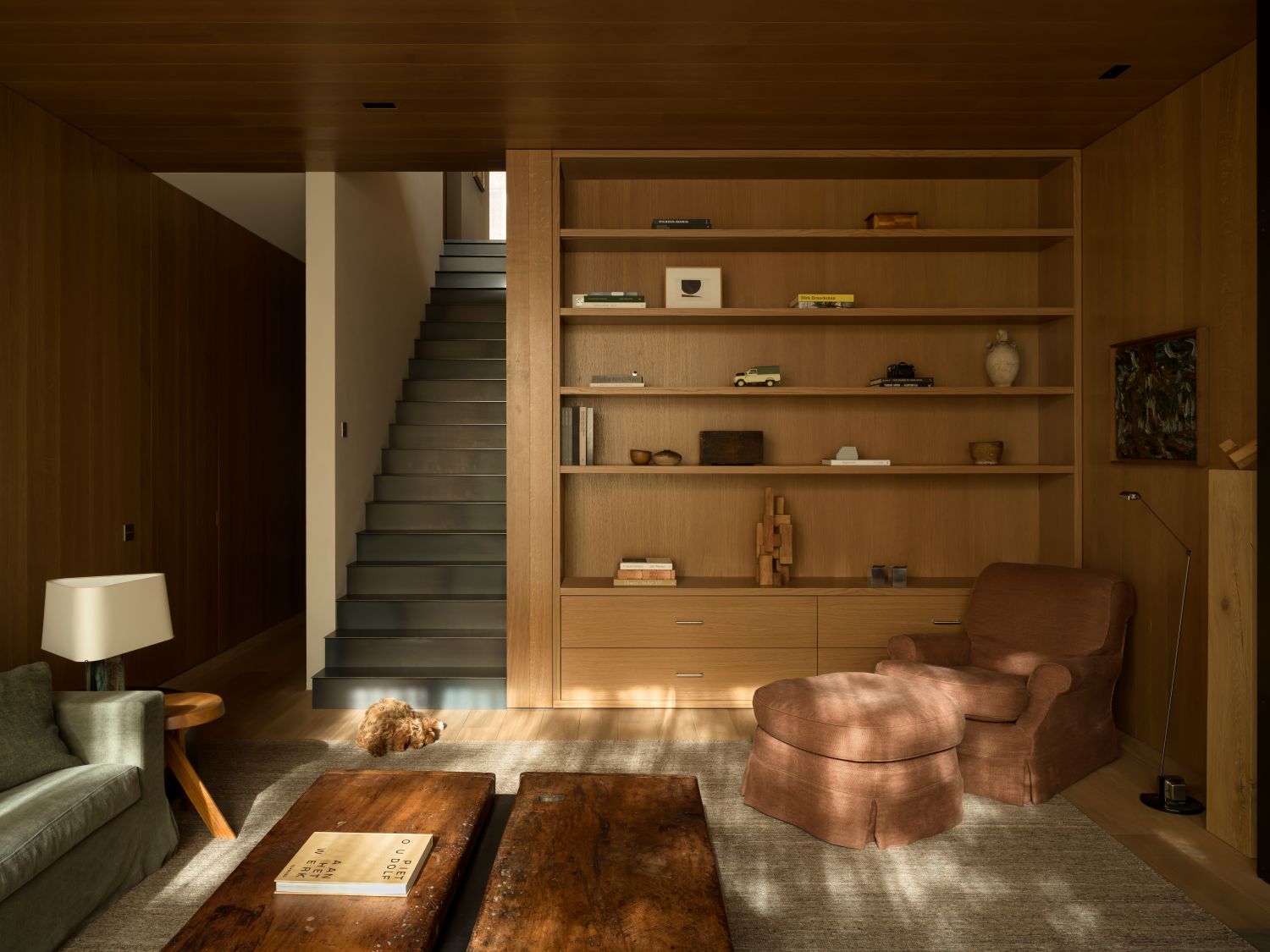 Warm modern living room with built-in oak shelving, a brown upholstered armchair with ottoman, rustic wooden coffee tables, and soft natural light highlighting minimalist decor.