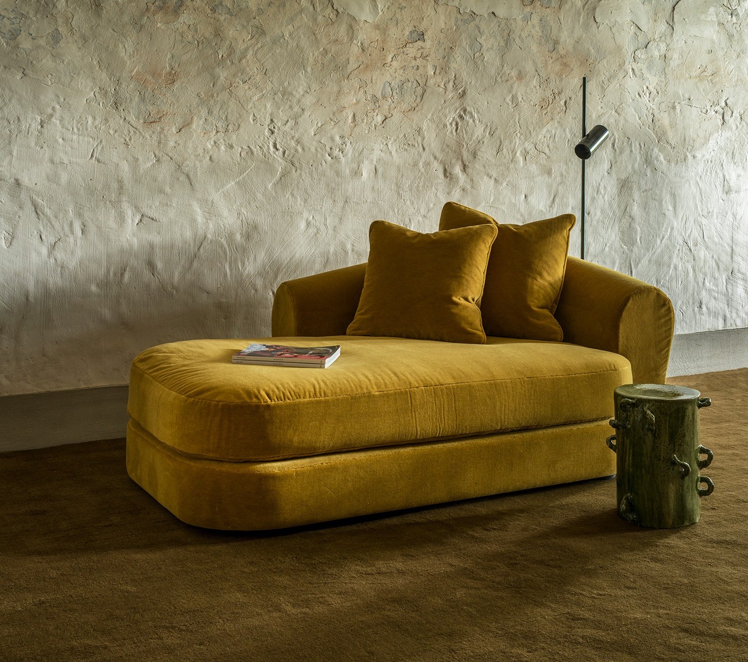 Sophia chaise longue in mustard velvet with two pillows, paired with a small wooden stool, set against a textured wall and soft brown carpet.