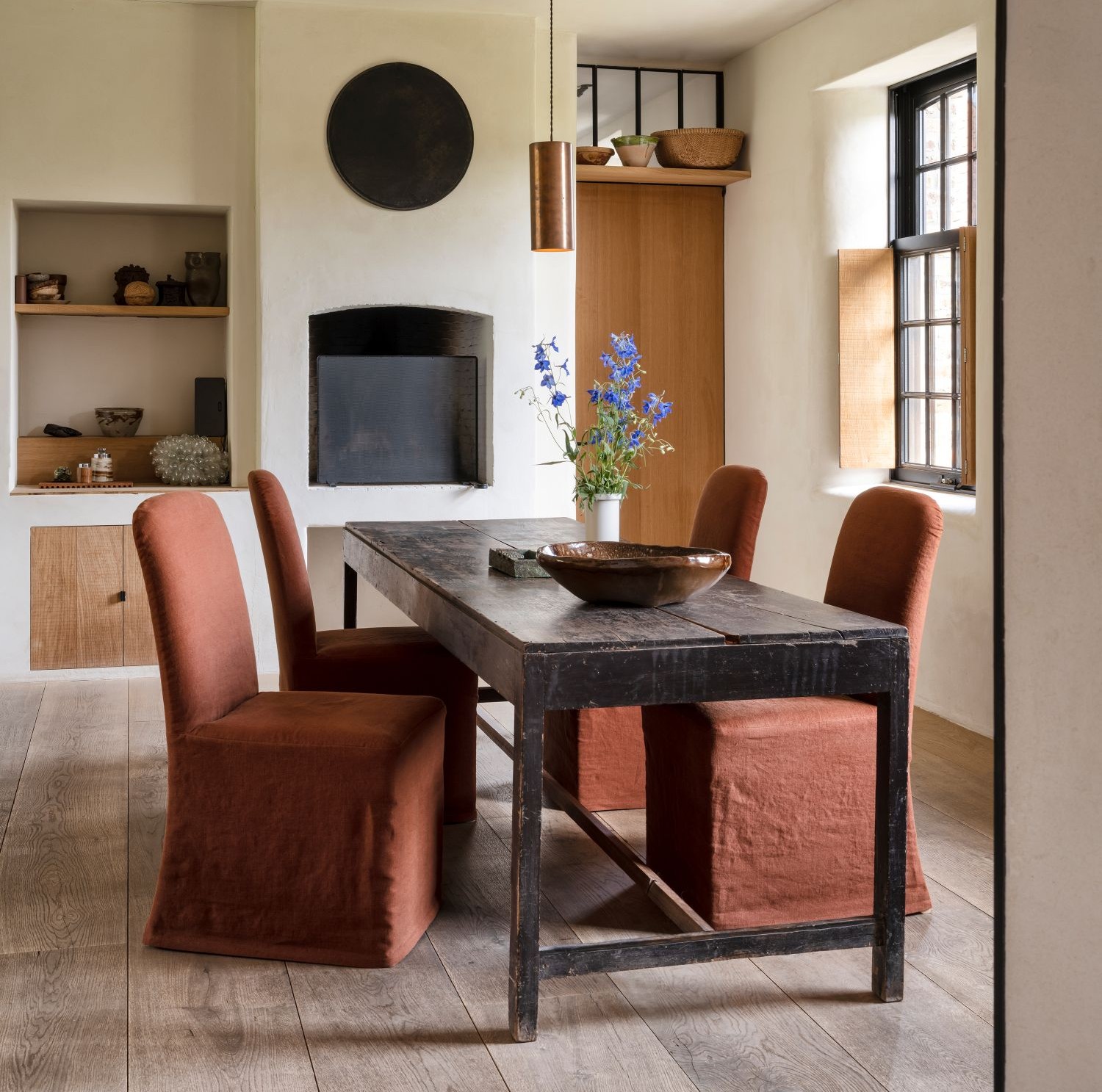 Dining room with rustic wooden table and four Julien dining chairs in rust-red slipcovers by Collett-Victor, styled with ceramic bowl and vase of blue flowers.