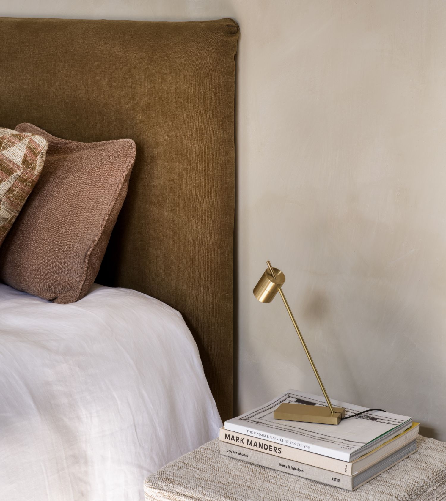 Mia headboard by Collett & Victor in a warm brown fabric, styled with pillows, white bedding, and a brass bedside lamp on a stack of books.