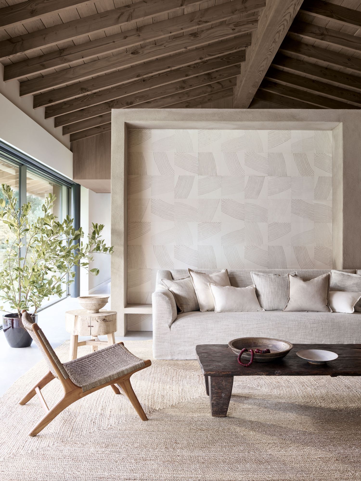 Bright living room featuring the Toscana fixed seat sofa by Collett & Victor in a neutral fabric, paired with natural wood furniture, woven textures, and a serene backdrop of soft geometric wall art under exposed wooden beams.