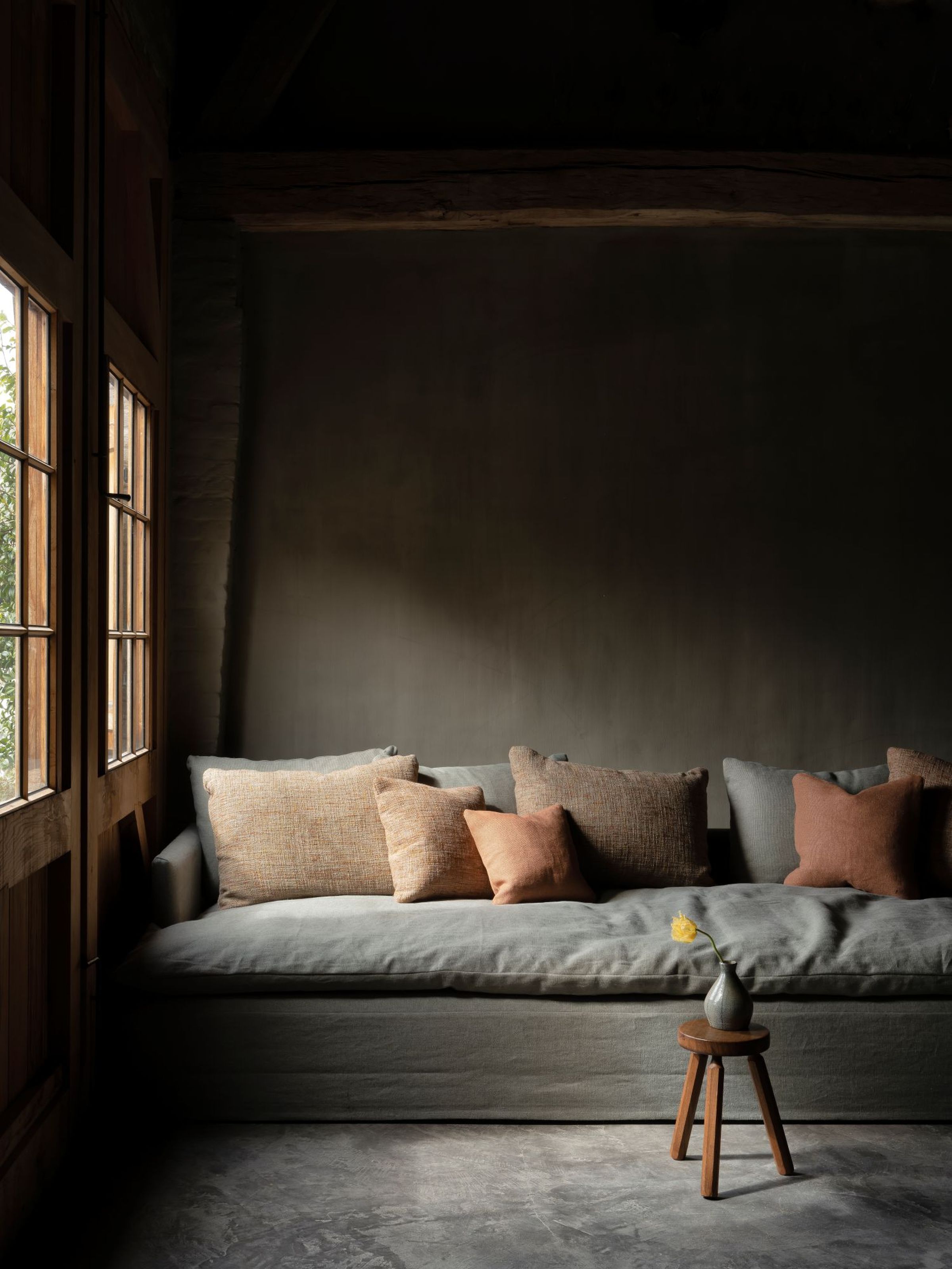 Bilbao sofa in soft grey fabric with deep seat and low arms, styled with textured neutral and terracotta cushions, placed in a dimly lit room beside wooden-framed windows and a small stool holding a single yellow flower in a ceramic vase.