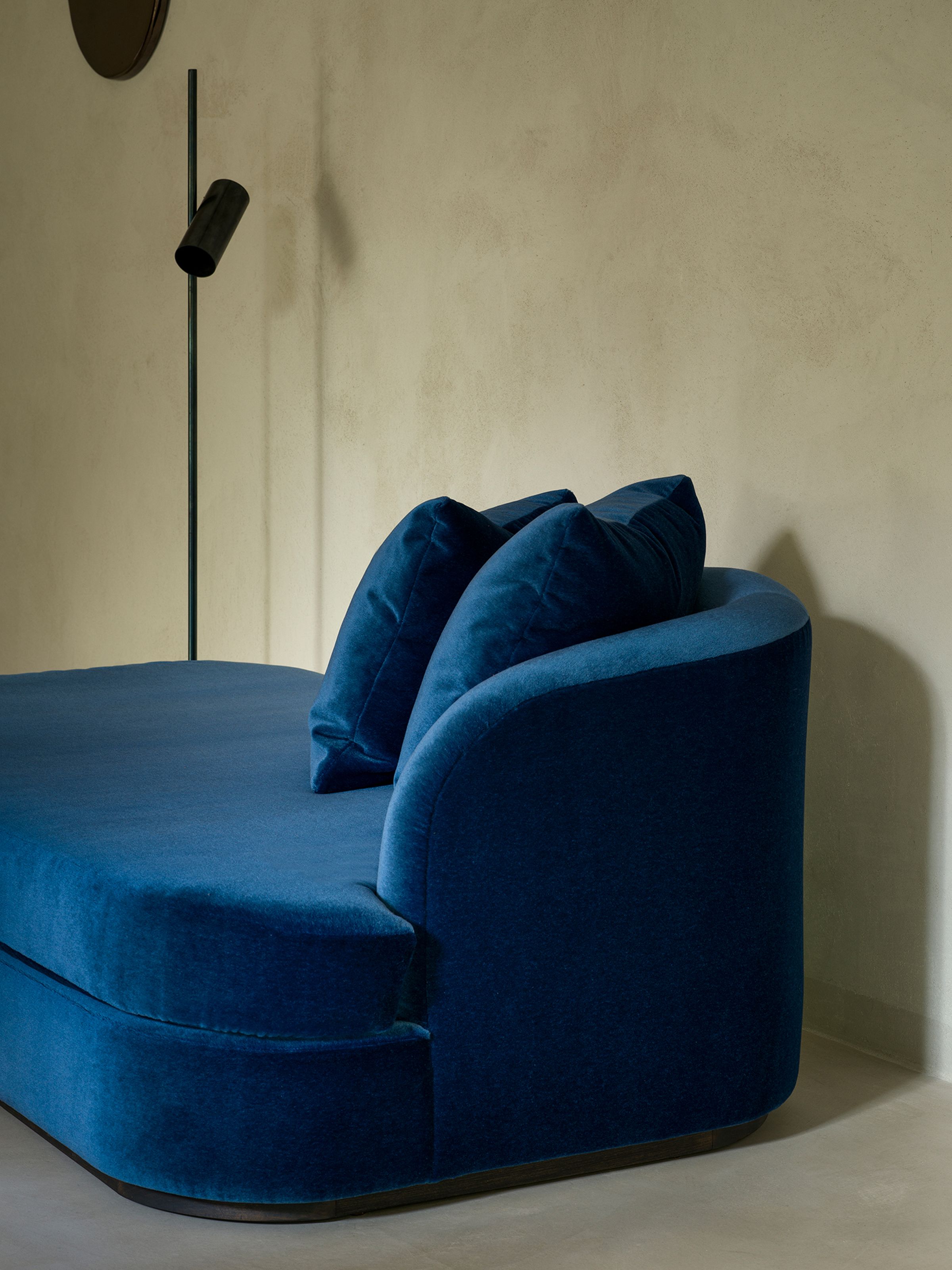 Close-up of a luxurious blue velvet chaise longue with two soft cushions resting on its curved backrest. A black floor lamp stands beside it, casting shadows on the smooth, neutral-colored wall.